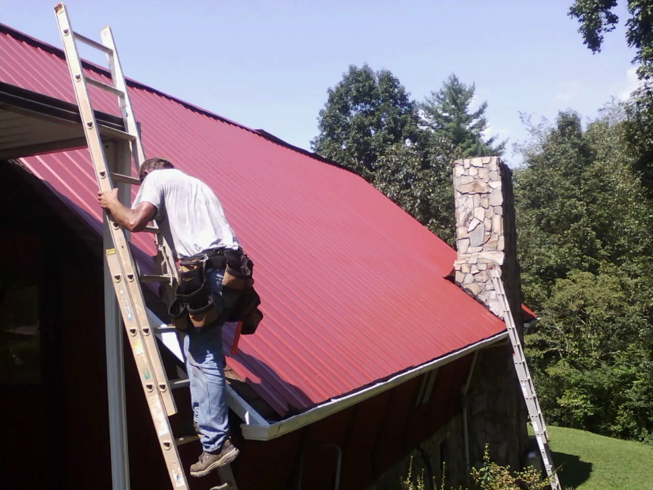 Roofer on a ladder beside a red metal roof and stone chimney — Soffit & Fascia Repair by Roofing Brothers in Lakehead